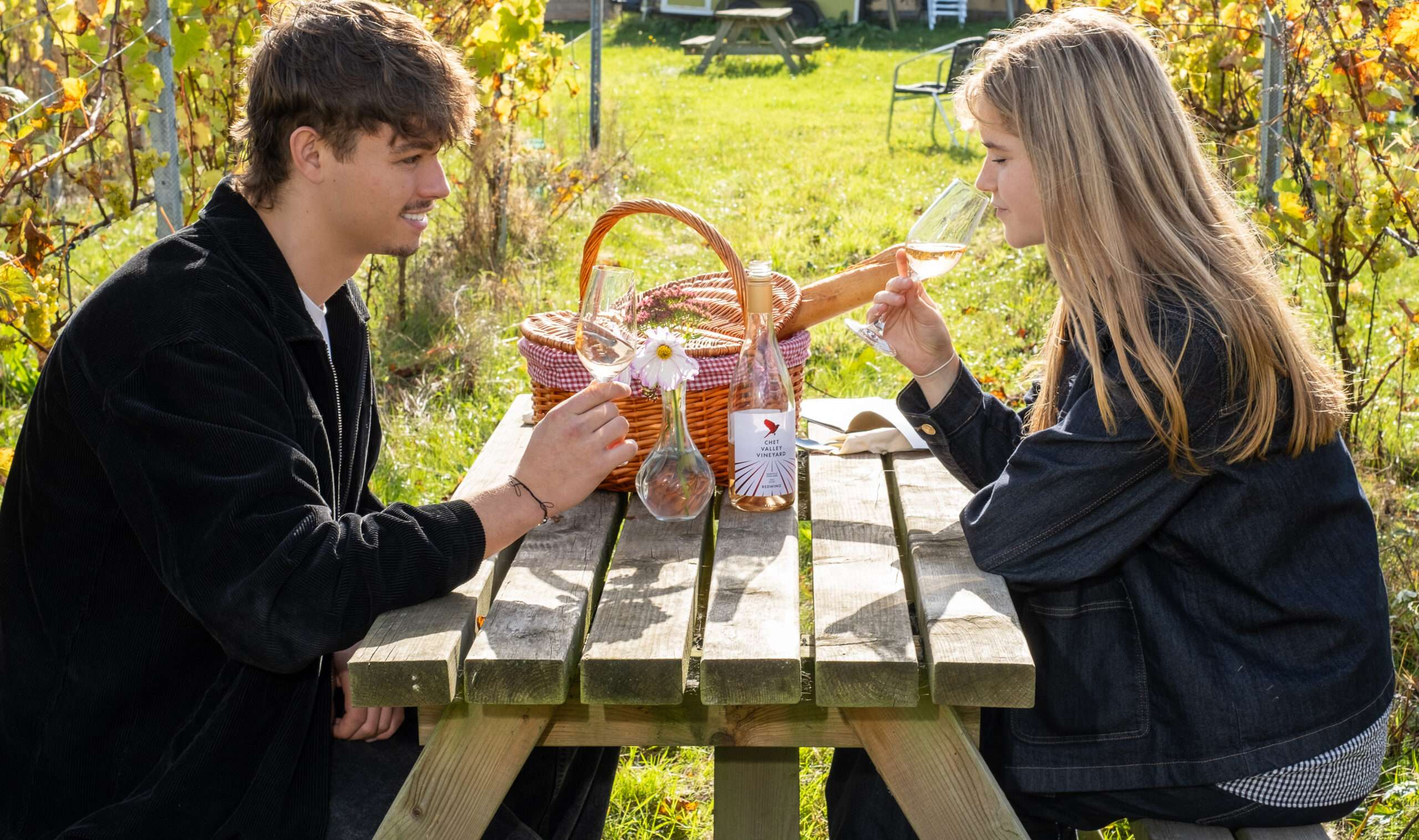 Visitors enjoying drinks and local produce from a picnic basket while seated outdoors overlooking the vineyard.