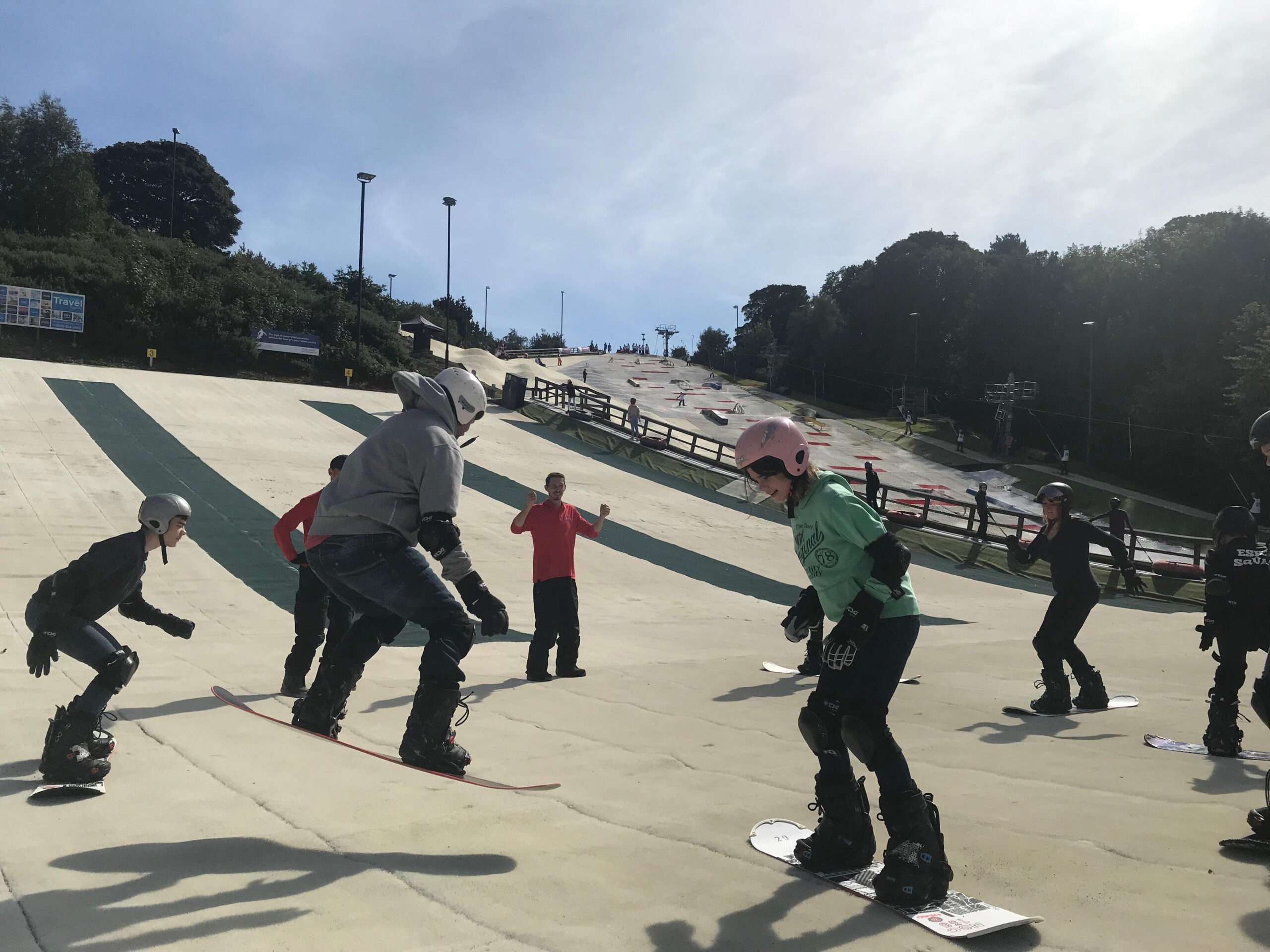 People receiving instruction on a dry ski slope, with learners practising on the beginner area and ski lift infrastructure visible in the background.