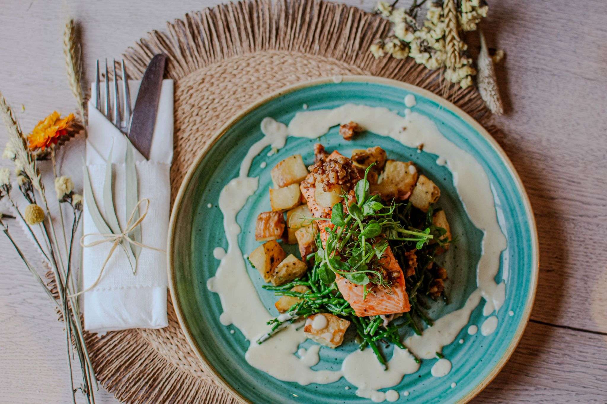 Close-up of a beautifully presented restaurant meal served on a turquoise plate, featuring fish, roasted potatoes, fresh greens and a creamy sauce.