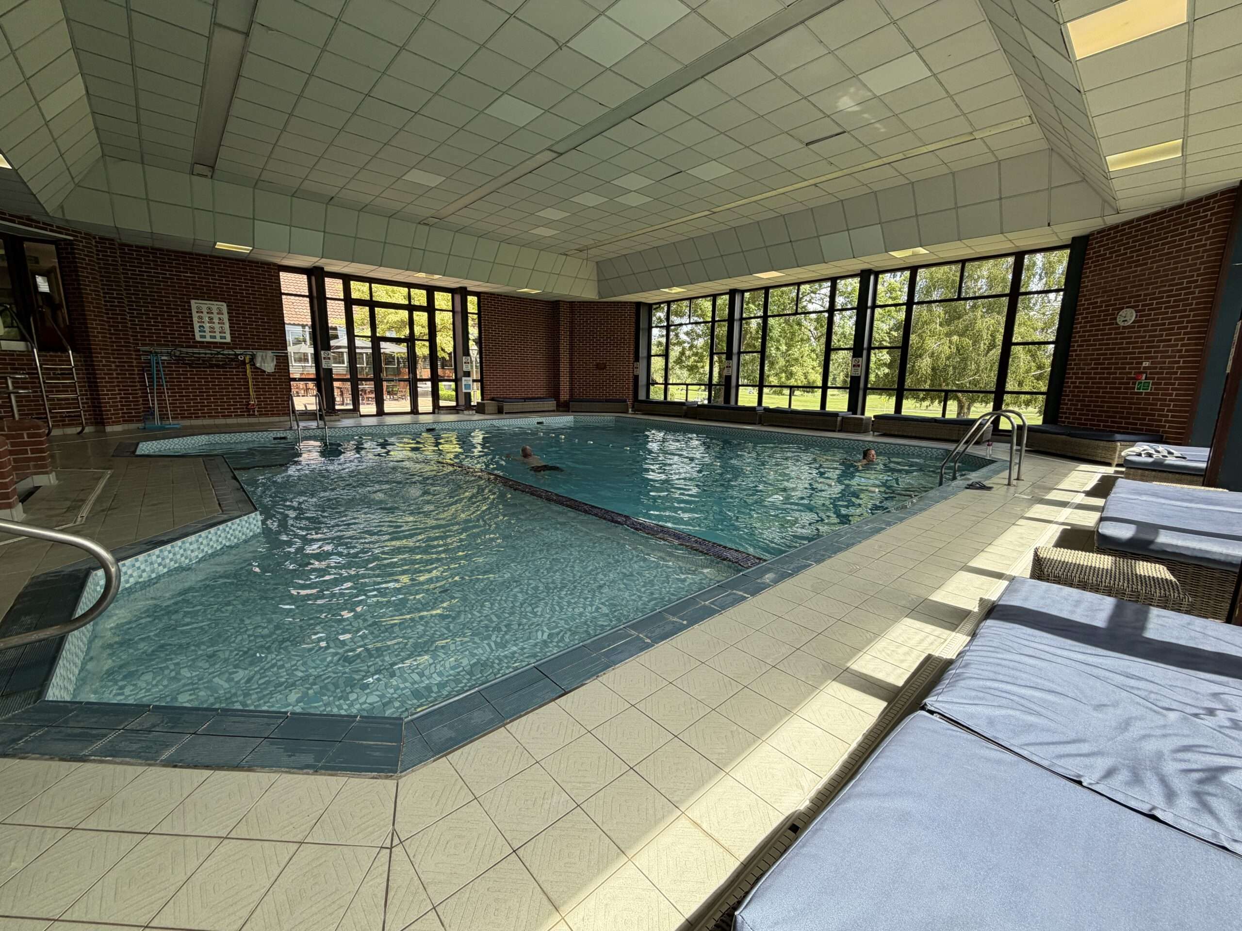 Indoor swimming pool with large windows overlooking trees and landscaped grounds.